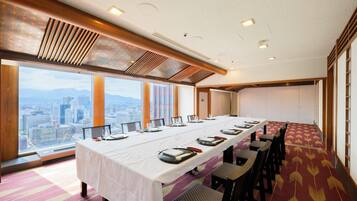 Un salón de conferencias con una larga mesa dispuesta para una reunión, amplias ventanas con vistas a la ciudad y una alfombra estampada.