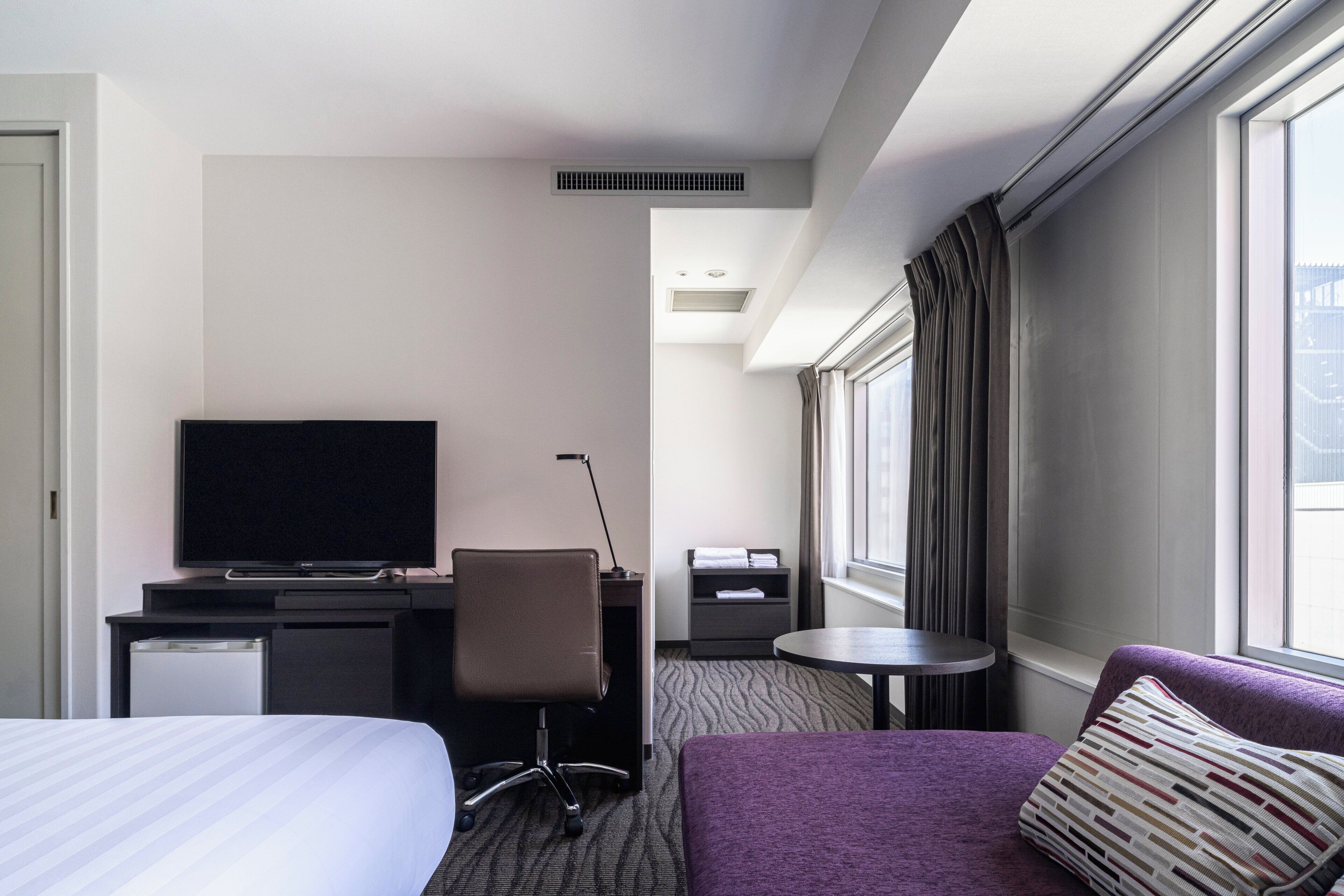 A hotel room with a bed, a desk with a television, a chair, a small table, and a window with curtains.