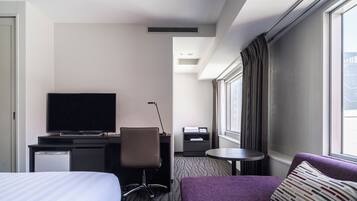 A hotel room with a bed, a desk with a television, a chair, a small table, and a window with curtains.