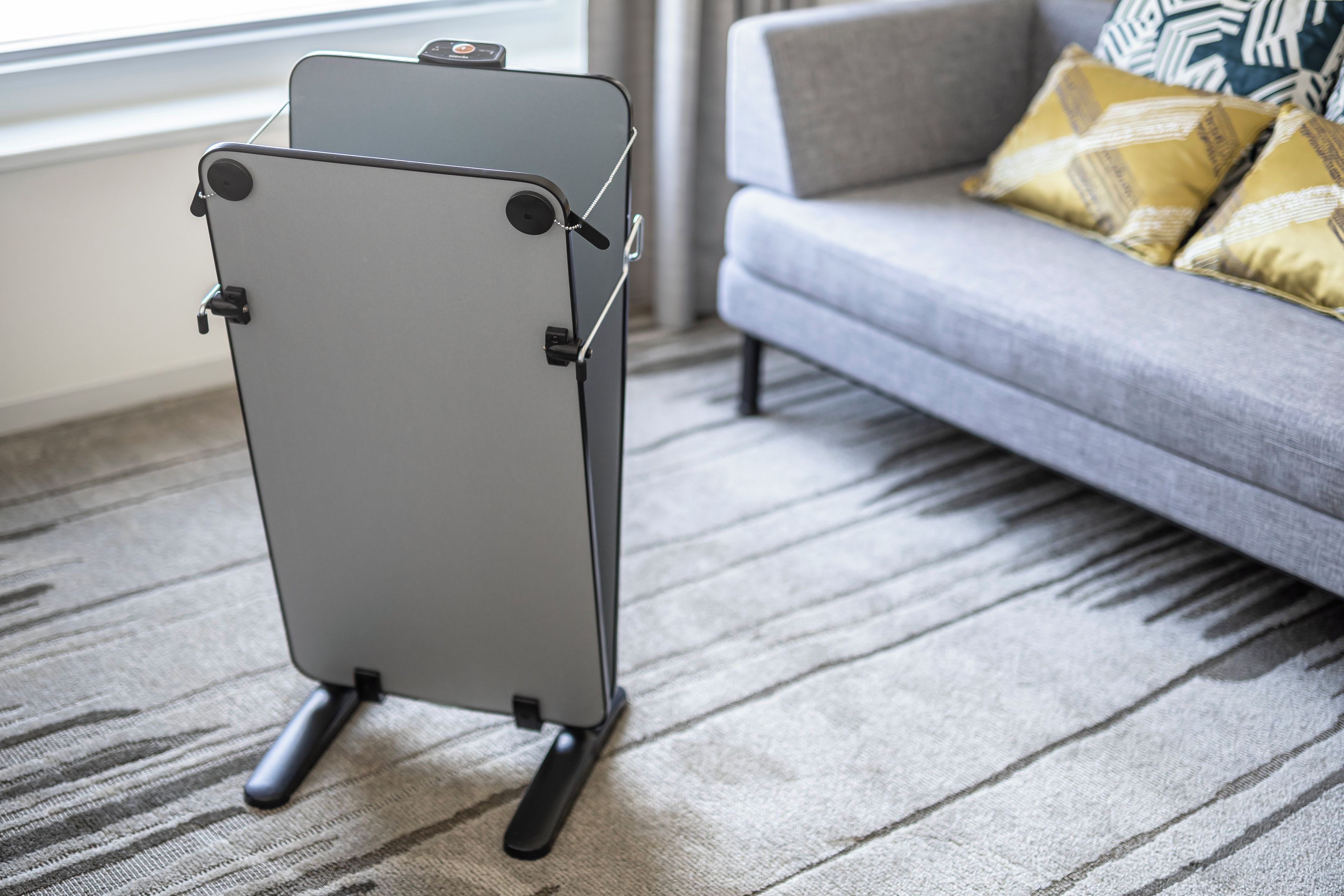 Una silla plegable gris con base negra, ubicada sobre un piso alfombrado en una habitación que tiene un sofá gris y cojines estampados al fondo.