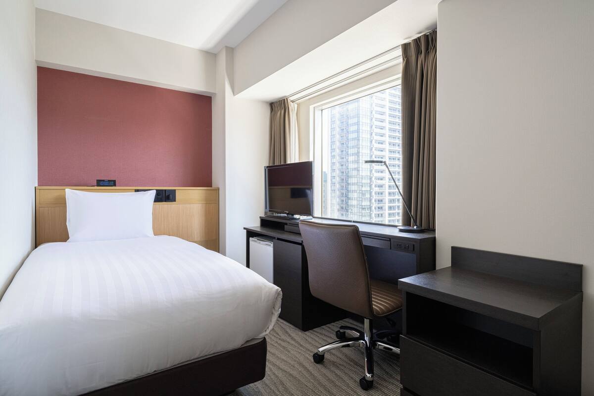 A hotel room with a bed, a desk, a chair, a TV, and a window with a city view.