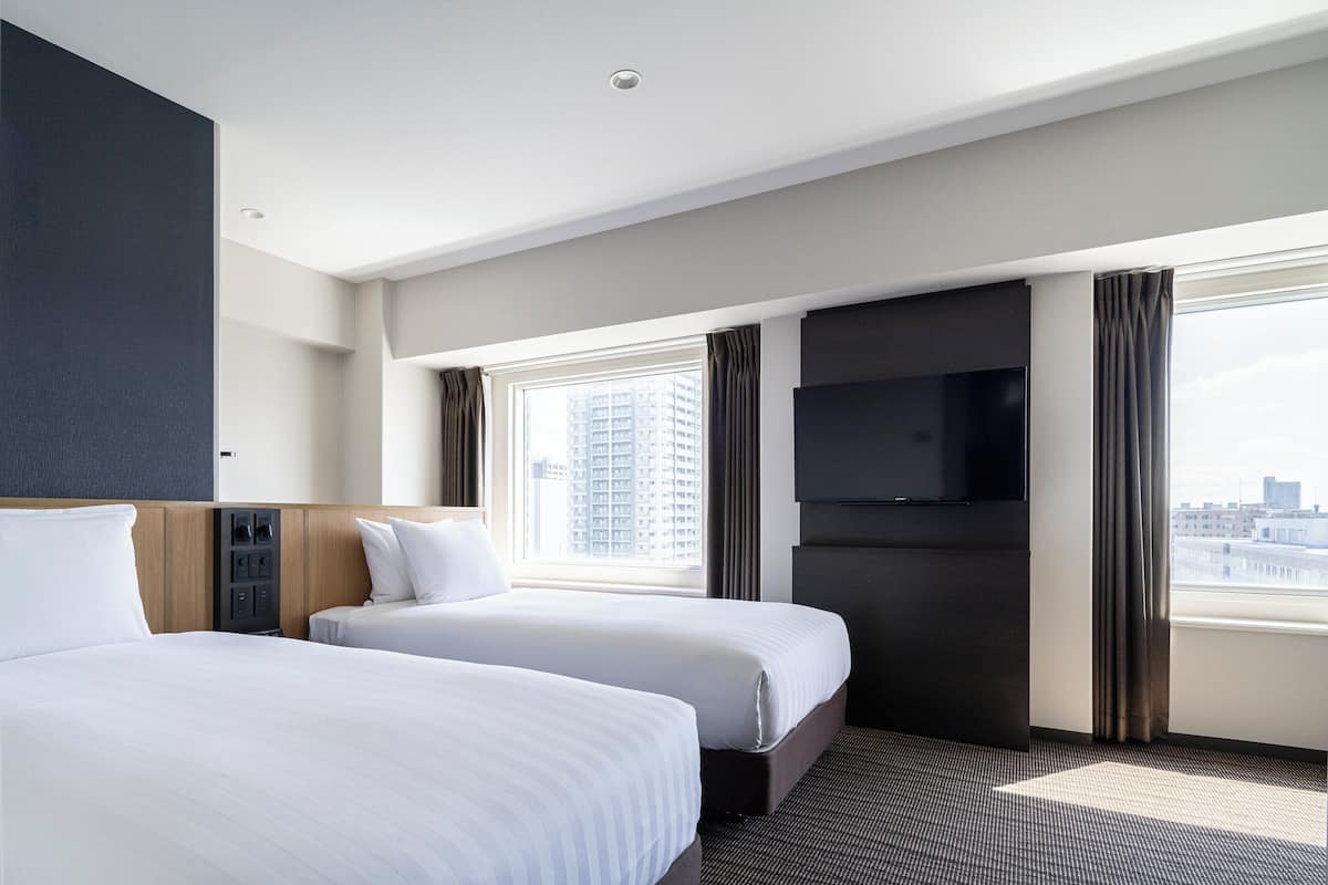 A hotel room with two beds, a flat-screen TV, a window with curtains, and a city view.