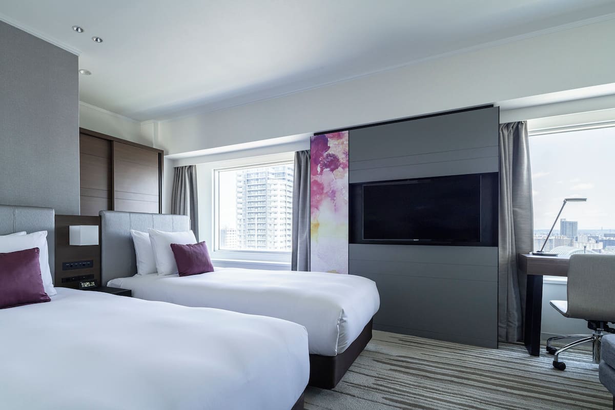 A hotel room with two beds, a desk, a TV, and a city view.