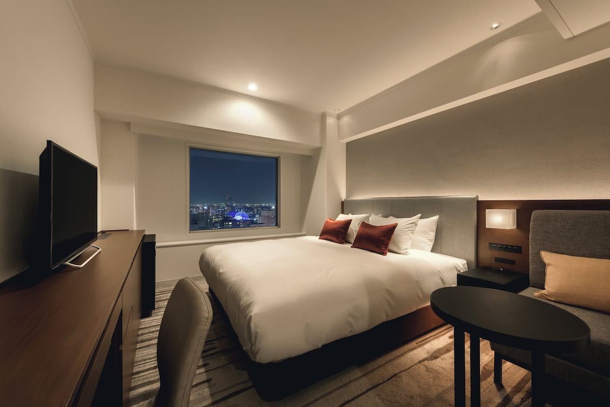 A modern hotel room with a large bed, a desk, a TV, and a window with a city view.