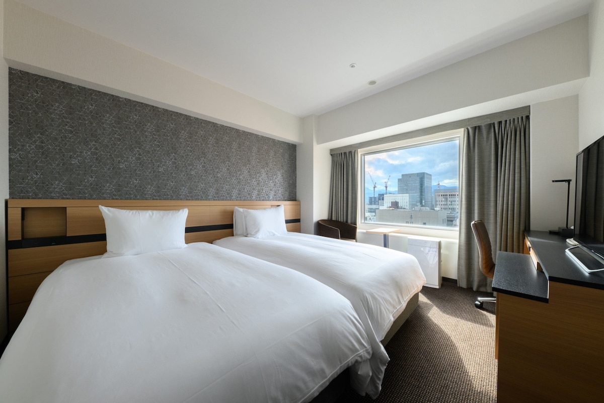 A hotel room with two beds, a desk, and a large window with a city view.