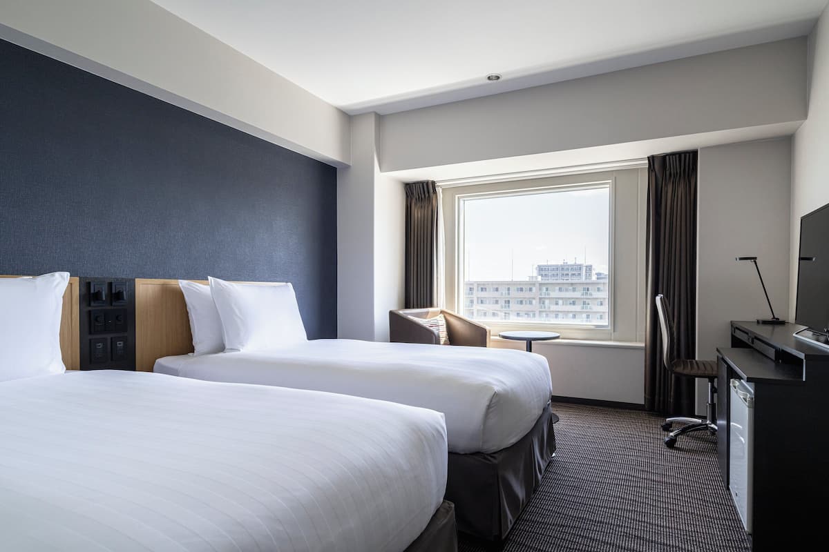 A hotel room with two beds, a desk, a chair, a small table, and a window with curtains.