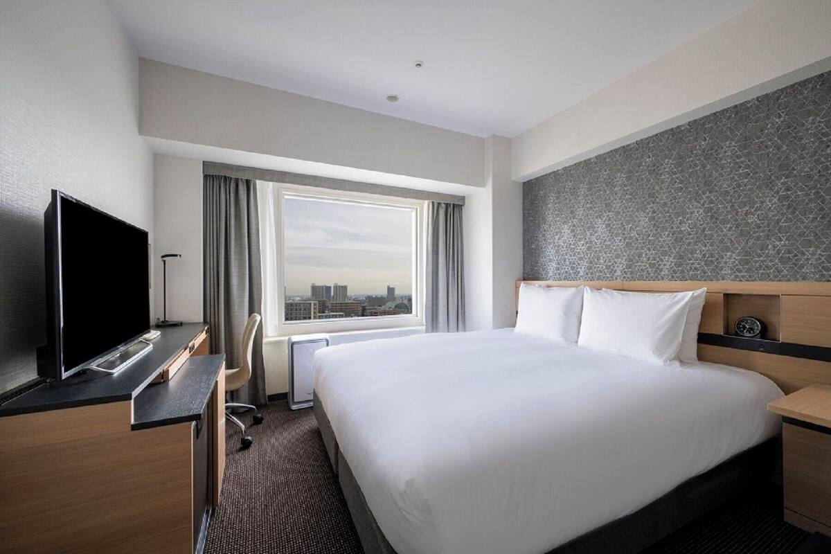 A hotel room with a large bed, a desk with a flat-screen TV, a chair, and a view of the city through a large window.