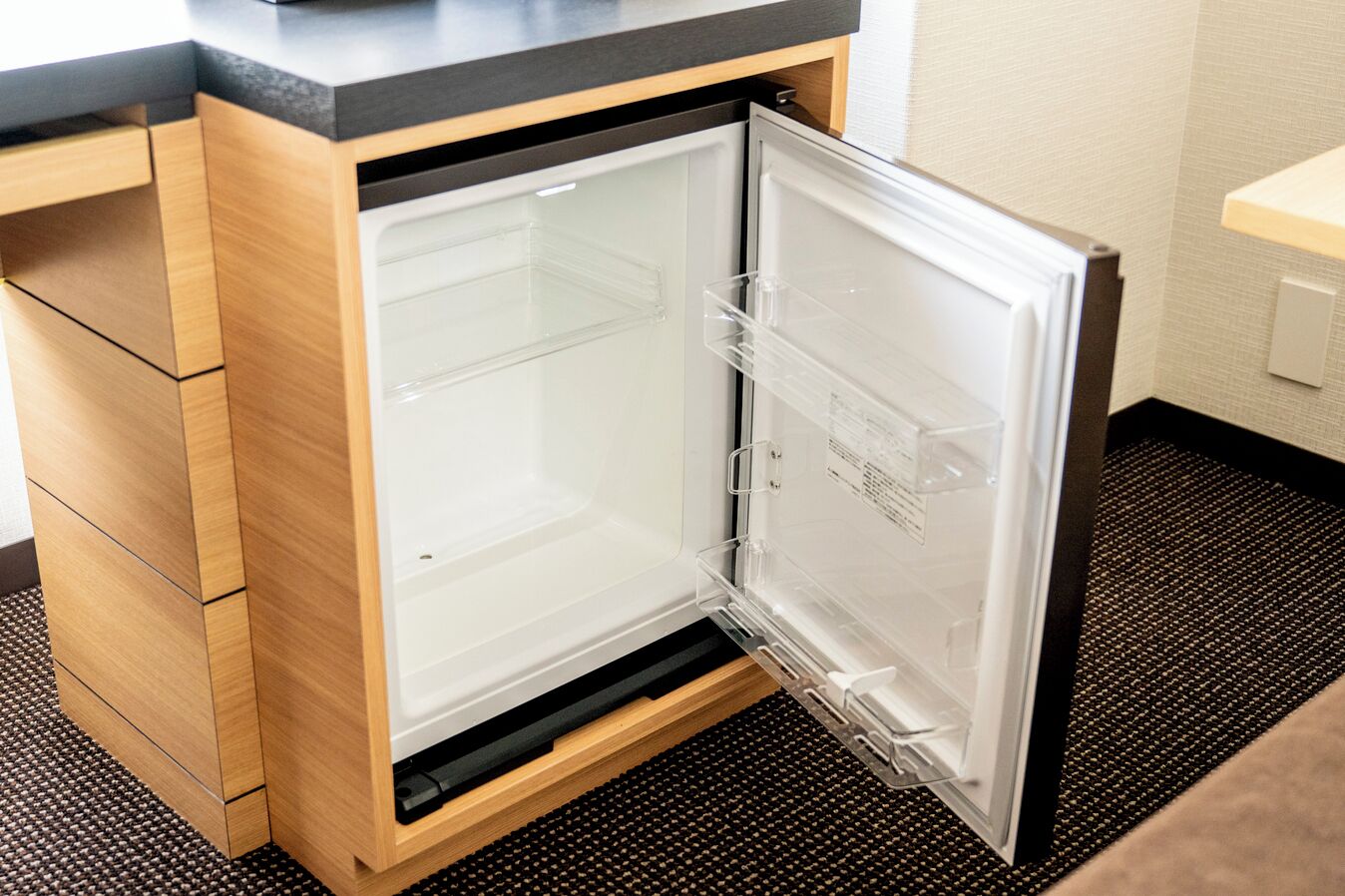 A compact refrigerator with a transparent door, built into a wooden cabinet with a black countertop above.