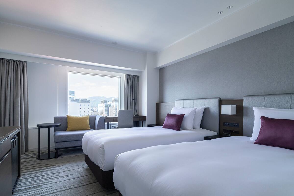 A hotel room with two beds, a desk, a chair, and a view of the city.