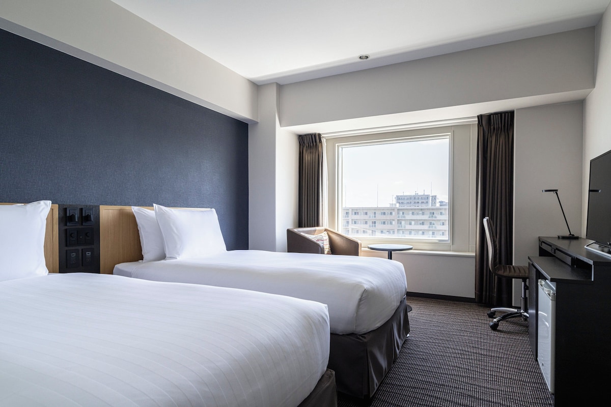 A hotel room with two beds, a desk, a chair, a small table, and a window with curtains.