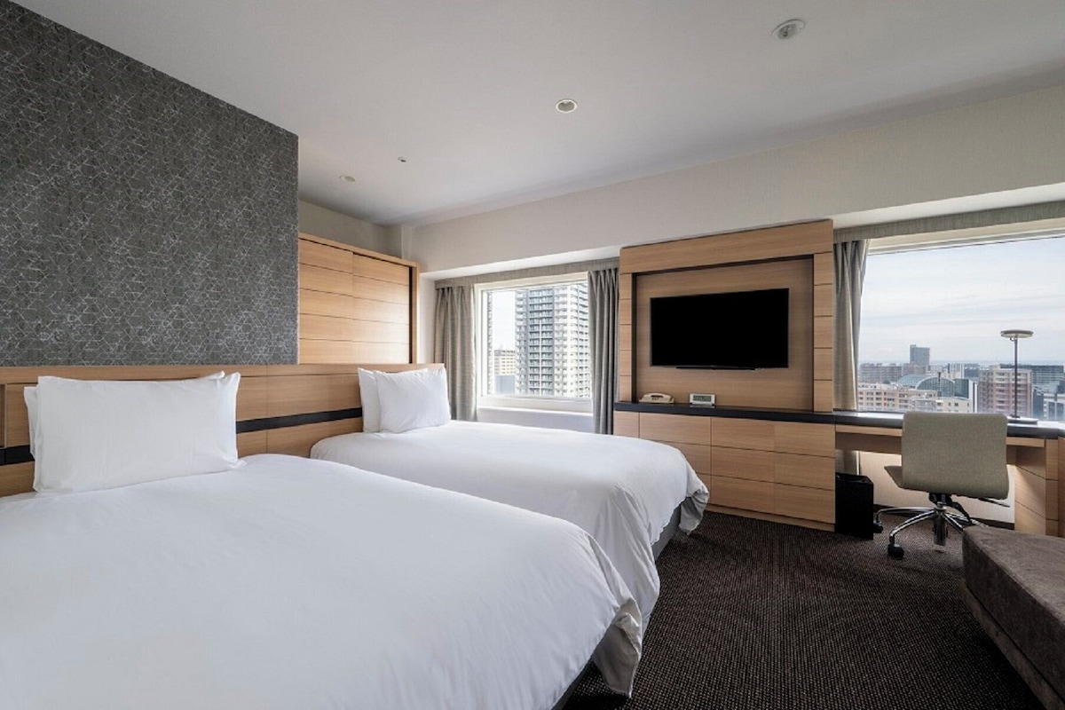 A hotel room with two beds, a TV, a desk, and a view of the city.