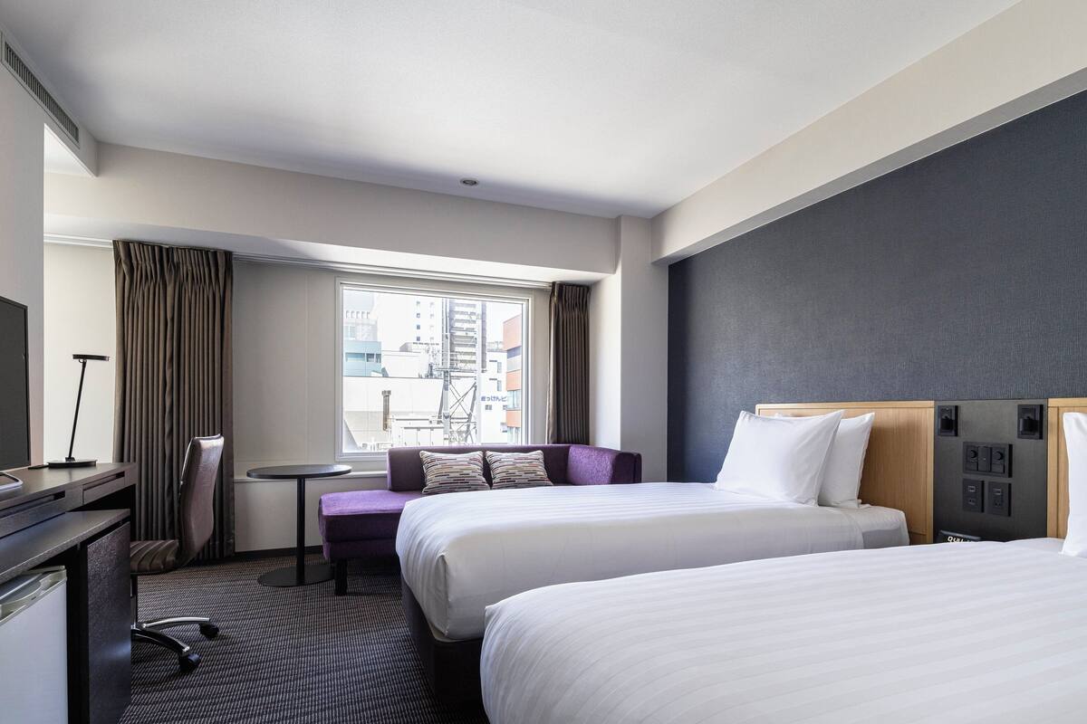 A hotel room with two beds, a desk, a chair, a small table, and a window with curtains.