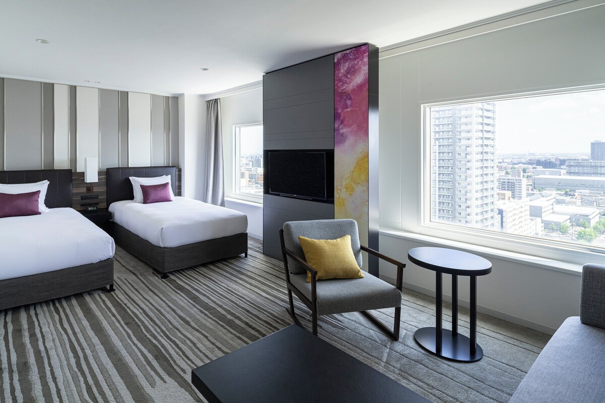 A hotel room with two beds, a TV, a chair, and a view of the city.