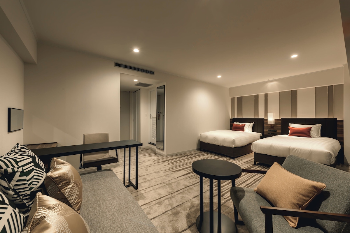 A modern hotel room with two beds, a sofa, a small table, and a mirror.
