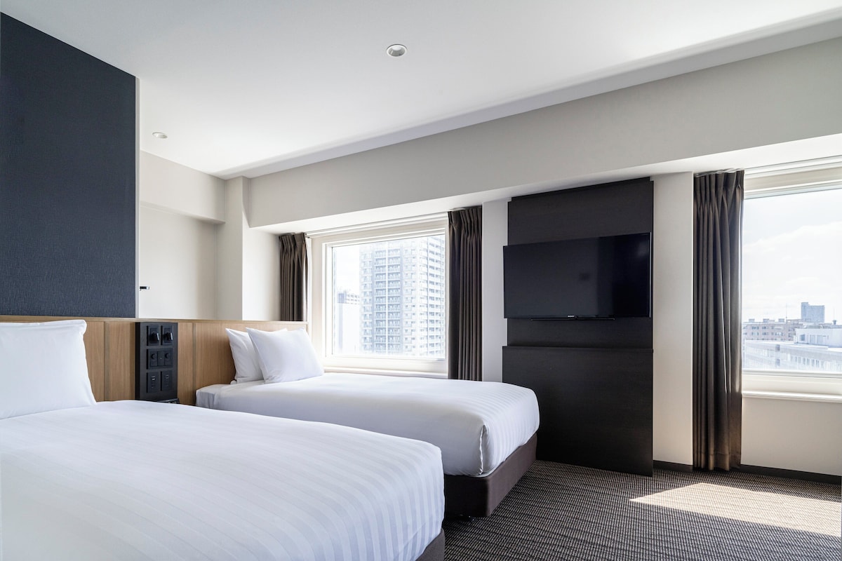 A hotel room with two beds, a flat-screen TV, a window with curtains, and a city view.