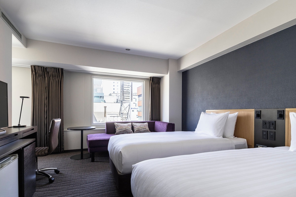 A hotel room with two beds, a desk, a chair, a small table, and a window with curtains.