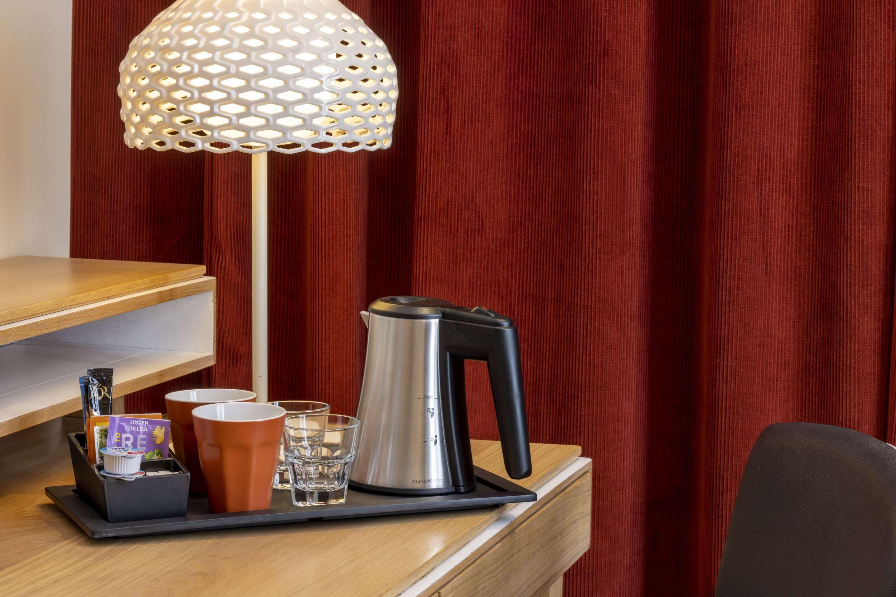 A hotel room with a wooden desk, a tray with a kettle, cups, glasses, and a decorative lamp.