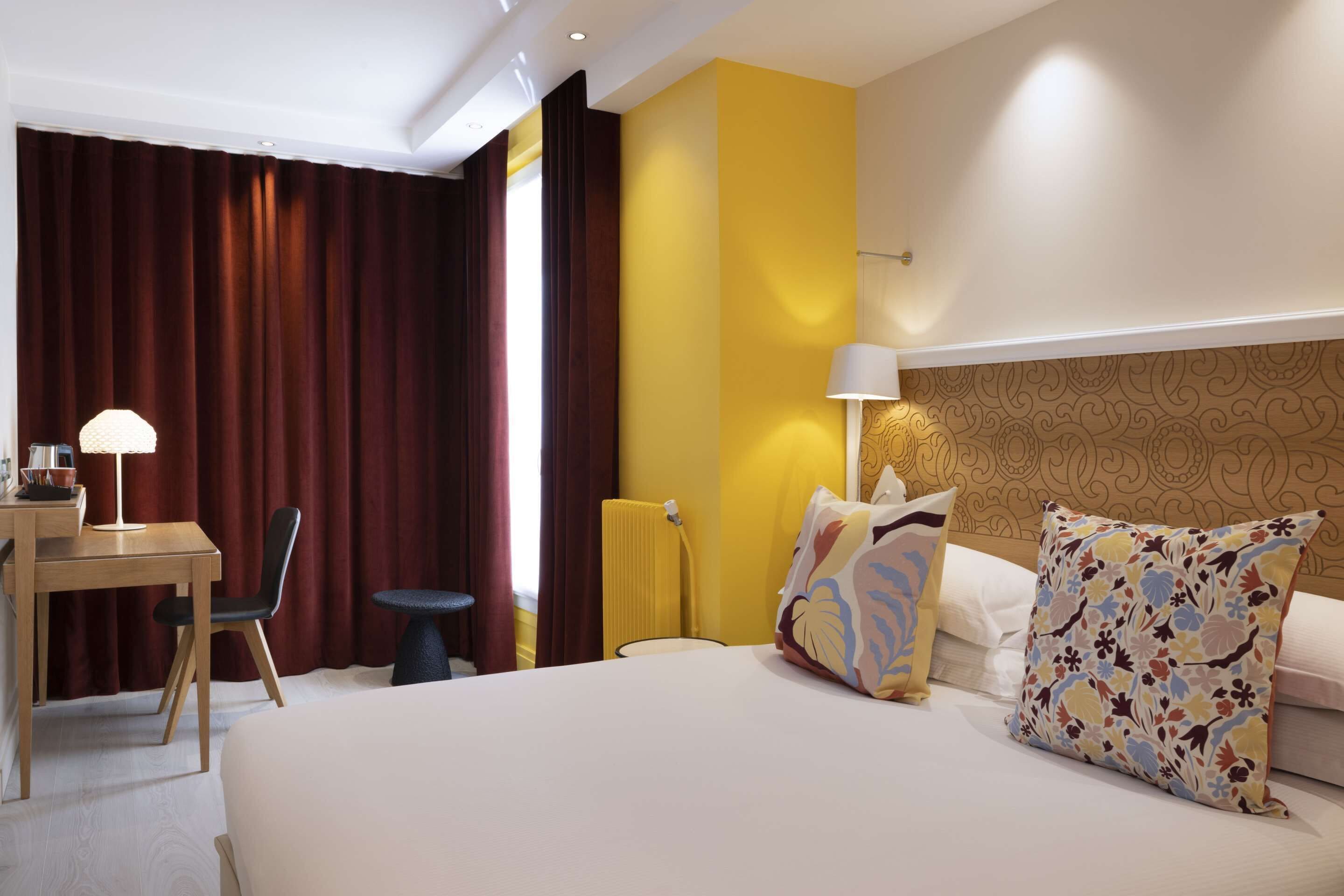 A hotel room with a bed, a desk, a chair, a small table, and a lamp.