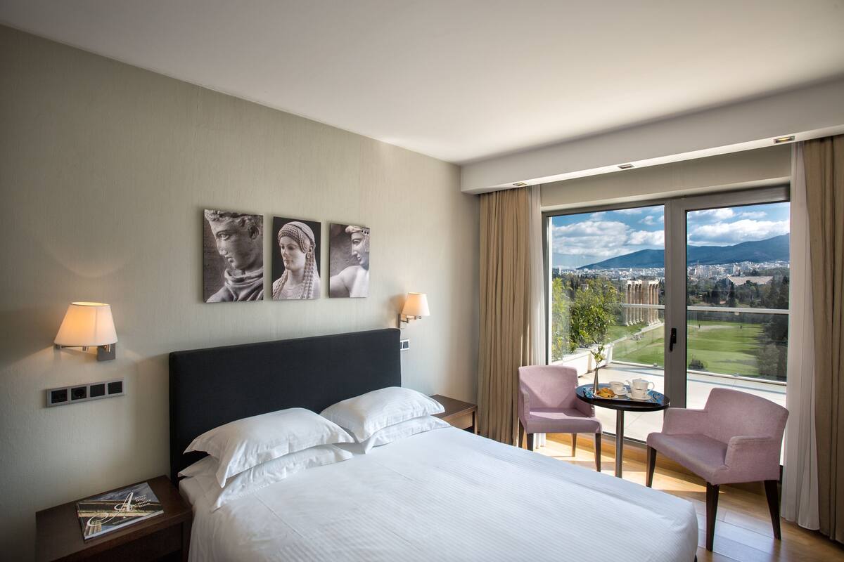 Camera d'albergo con un letto, due comodini, una sedia, un tavolino con tazza e piattino, e un'ampia finestra vista città e montagne.