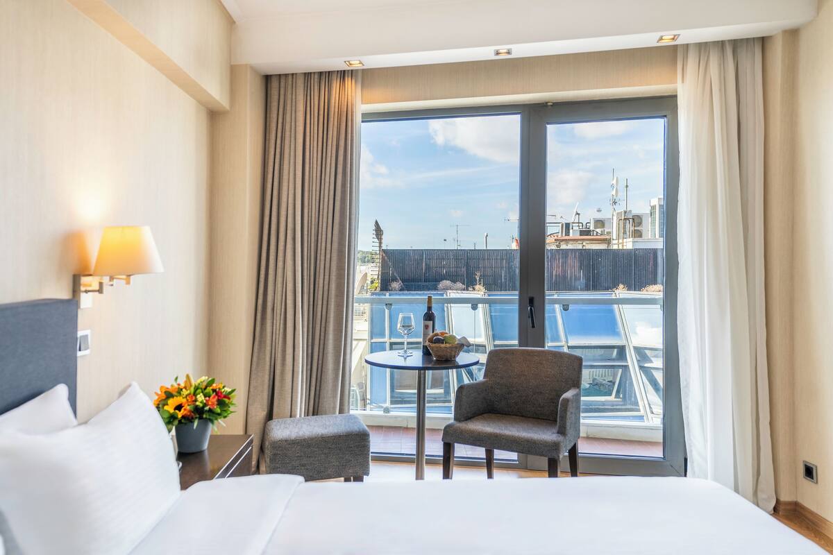 Una camera d'albergo con un letto, un tavolino con una bottiglia e della frutta, una sedia e un balcone con vista sulla città.