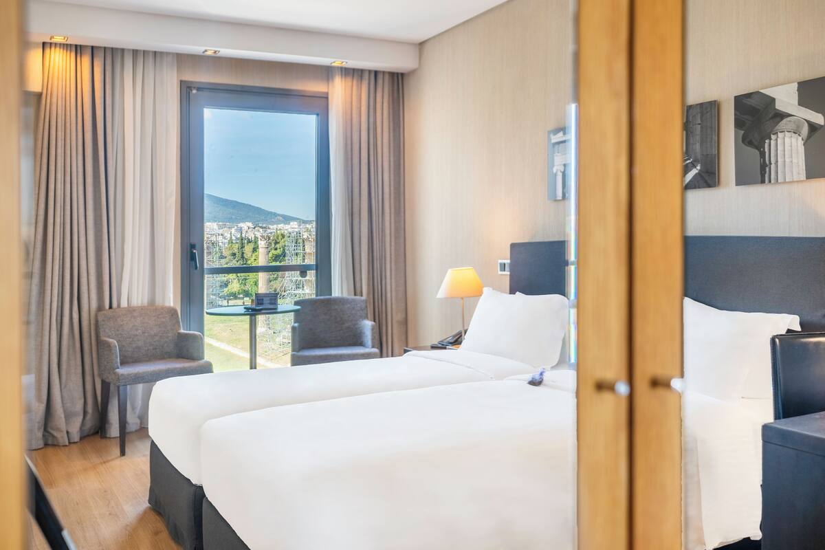 Una camera d'albergo con un letto grande, due poltrone, un tavolino, una lampada e vista sulla città attraverso un'ampia finestra.