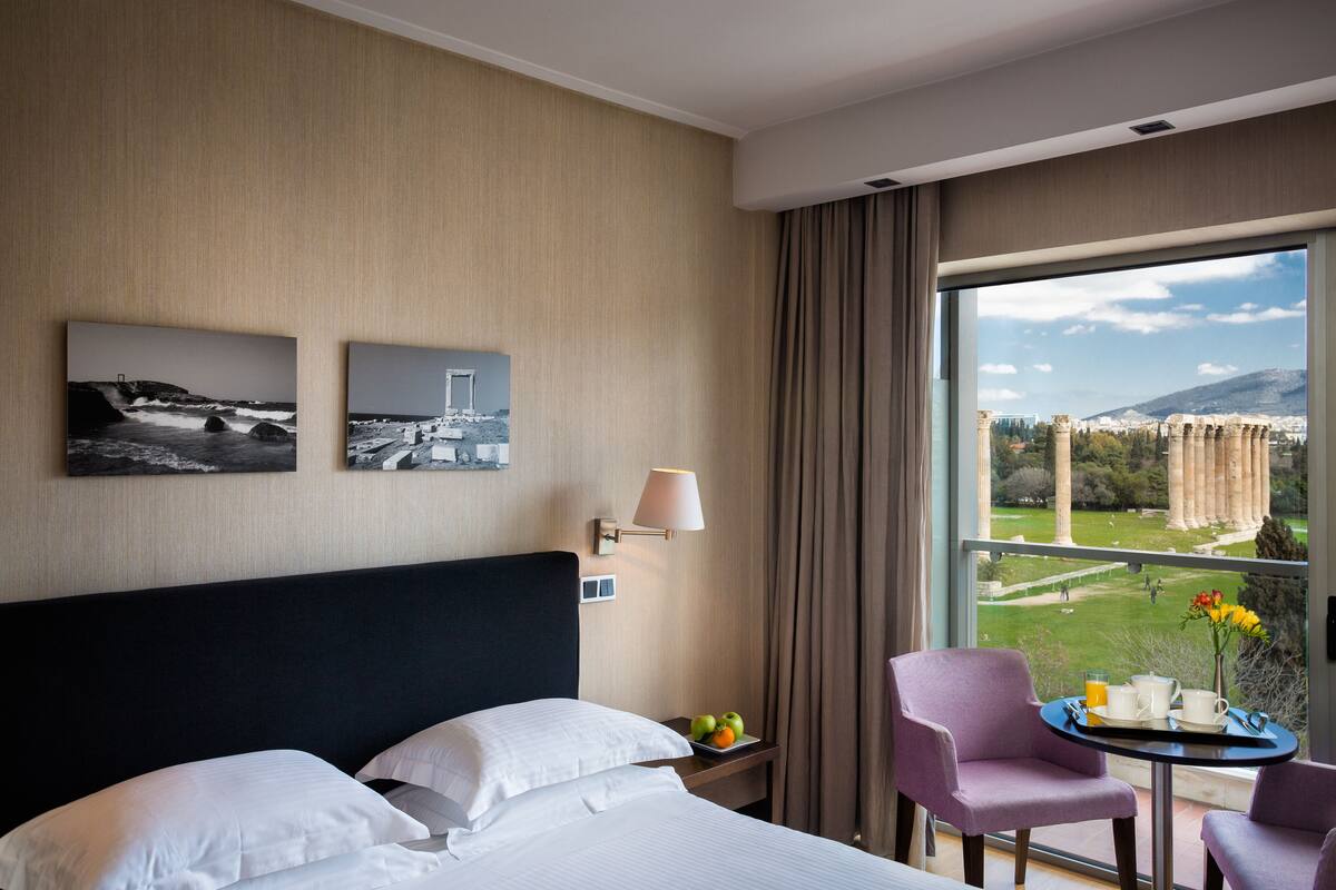 Una camera d'hotel con un letto grande, due poltrone, un tavolino con cibi per la colazione e vista sulla città e sulle montagne.