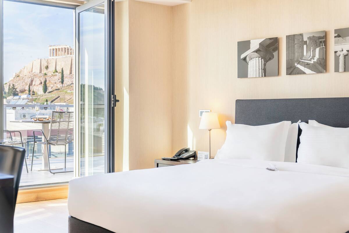 Camera d'albergo con un letto grande, un comodino con una lampada, un telefono e vista su rovine antiche tramite una porta in vetro.