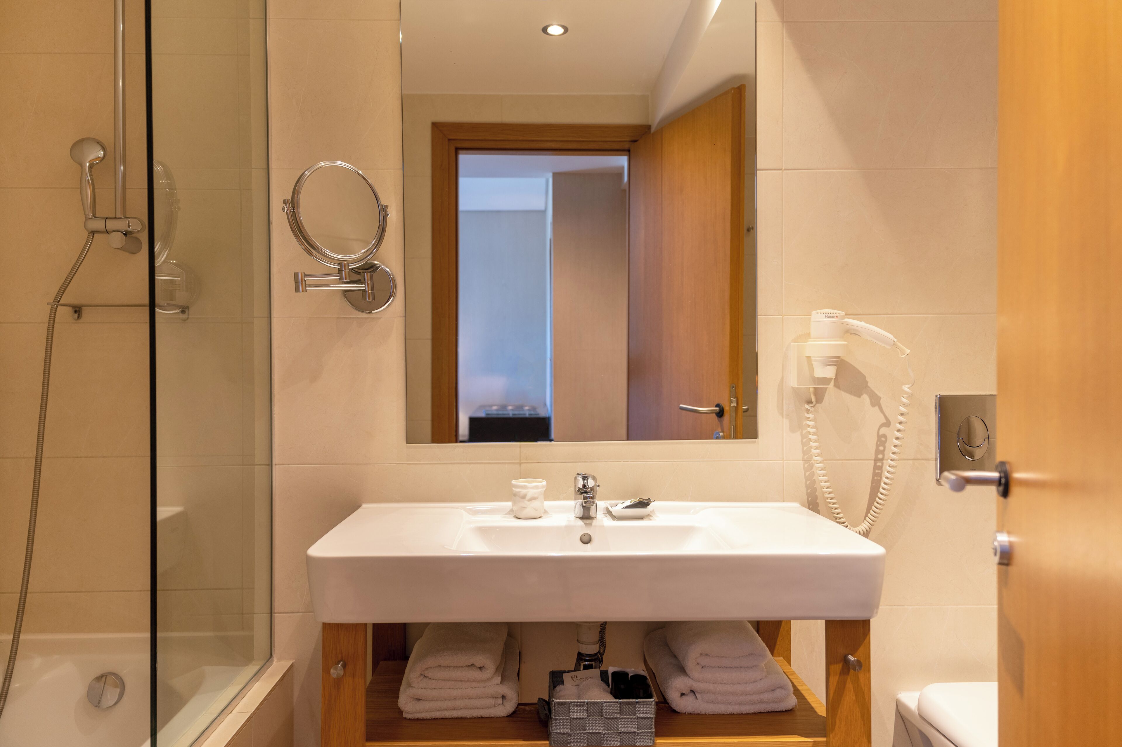 Een moderne badkamer met een douche met glazen wand, een wit wastafelmeubel met spiegel erboven, en een houten kastje met handdoeken.