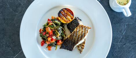 Un plato blanco con pescado a la parrilla, una rodaja de limón asado y una ensalada como acompañamiento sobre una superficie de piedra oscura.