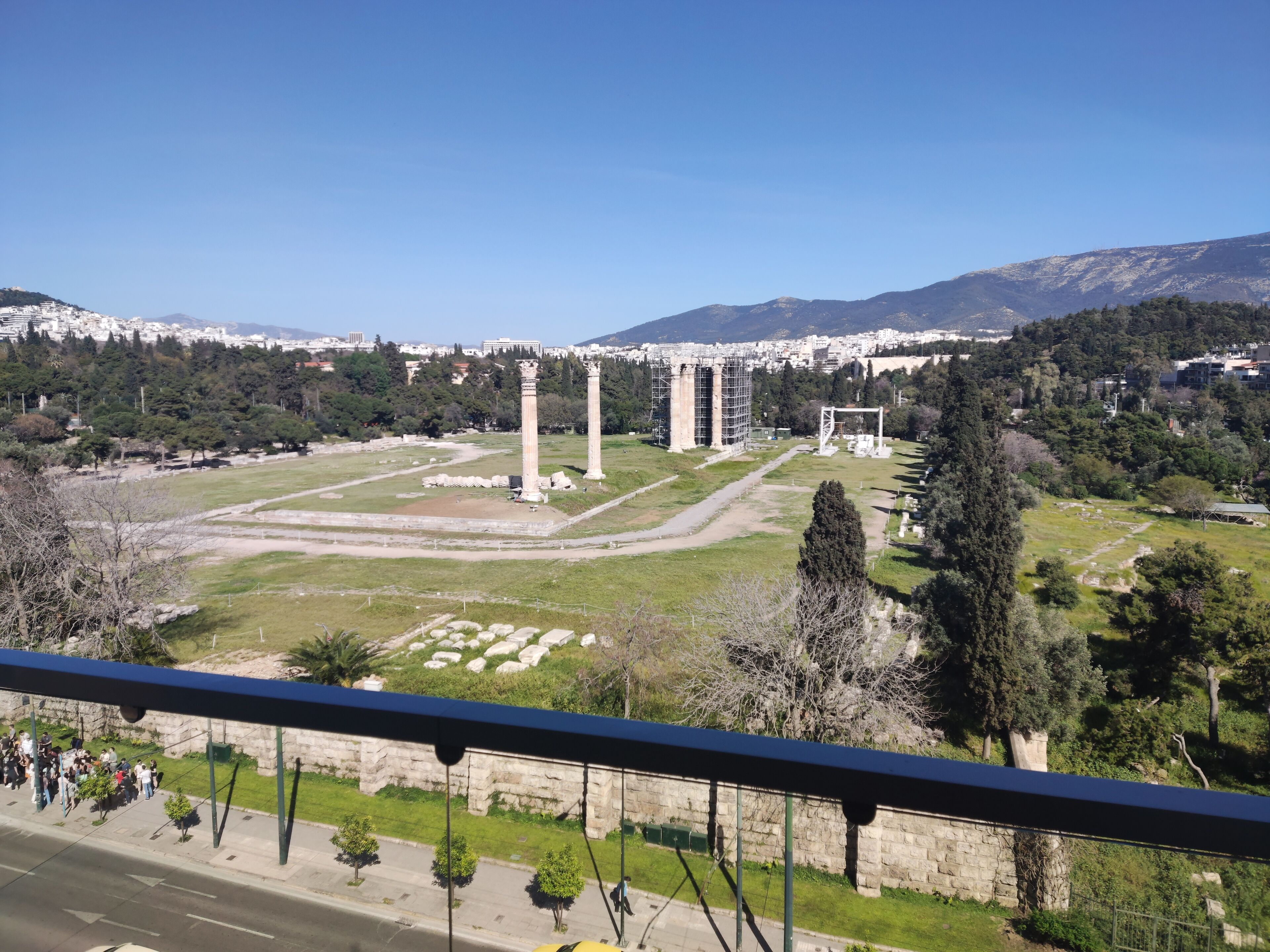 A view of ancient ruins with columns and a large open area, surrounded by trees and a cityscape in the background.