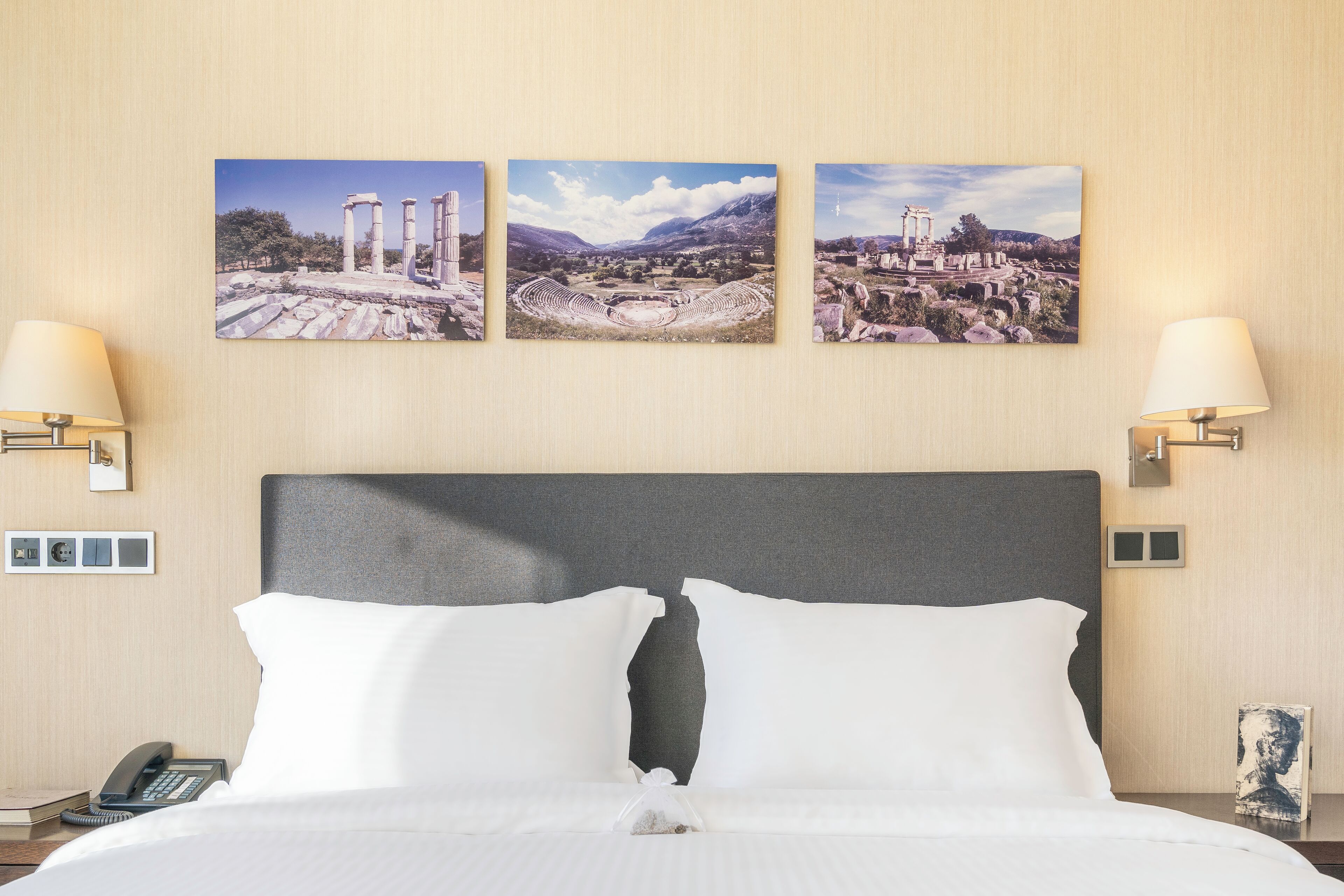 A hotel room with a large bed, two wall-mounted lamps, and three framed pictures depicting ancient ruins.