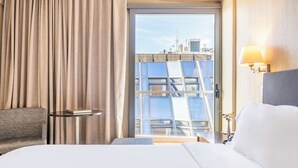 A hotel room with a bed, a small table, a chair, a window with a view of buildings, and a lamp.