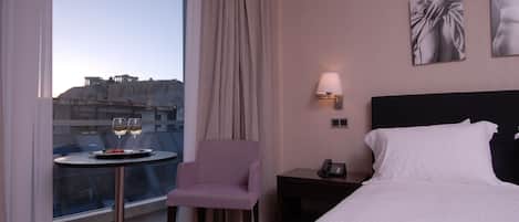 A hotel room with a bed, a chair, a small table with a plate and glasses, a nightstand with a phone, and a window with a view of ancient ruins.
