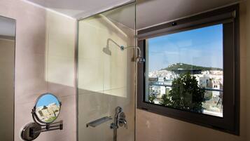 A bathroom with a glass shower enclosure, a window with a view of a cityscape, and a small mirror.