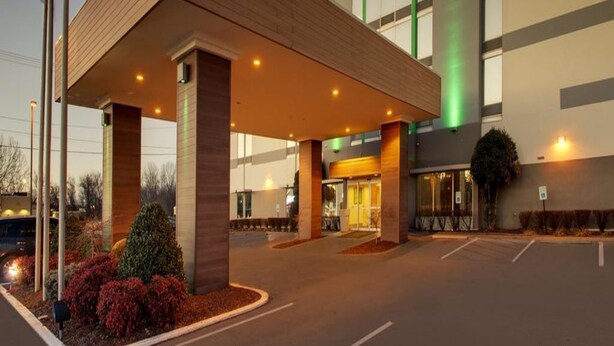 A modern hotel entrance with a covered walkway, a parking lot, and a flagpole.