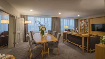 A hotel room with a dining table, chairs, a minibar, a TV, and a large window with curtains.