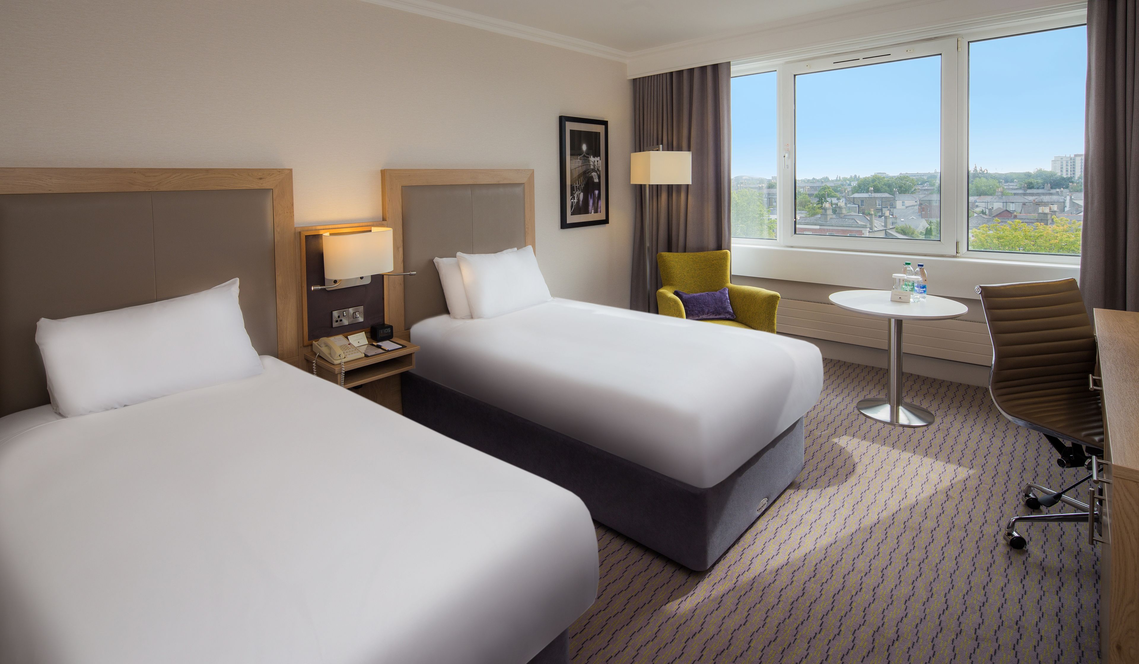 A hotel room with two beds, a desk, a chair, a small table, and a window with a city view.