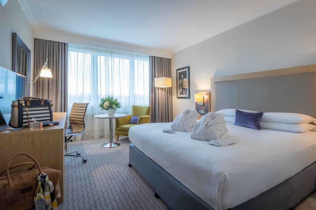 A hotel room with a large bed, a desk with a lamp, a chair, a small table with a plant, and a window with curtains.