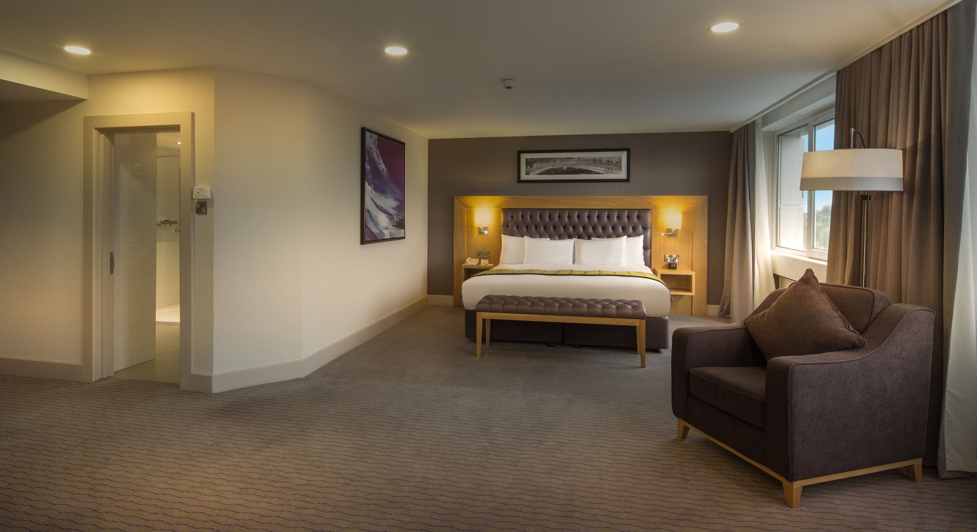 A hotel room with a large bed, a chair, a bedside table, and a window with curtains.