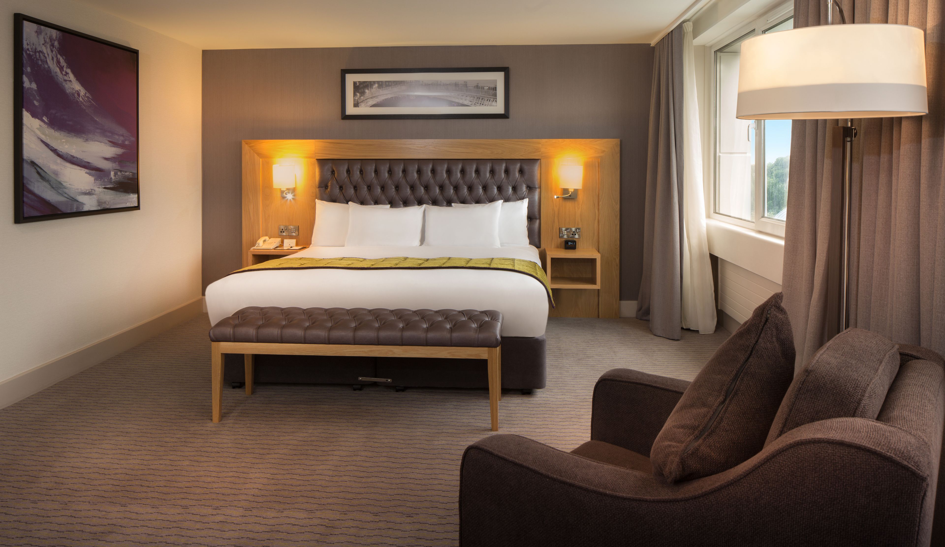 Une chambre d’hôtel avec un grand lit, une table de chevet avec une lampe, une chaise et un tableau encadré au mur.