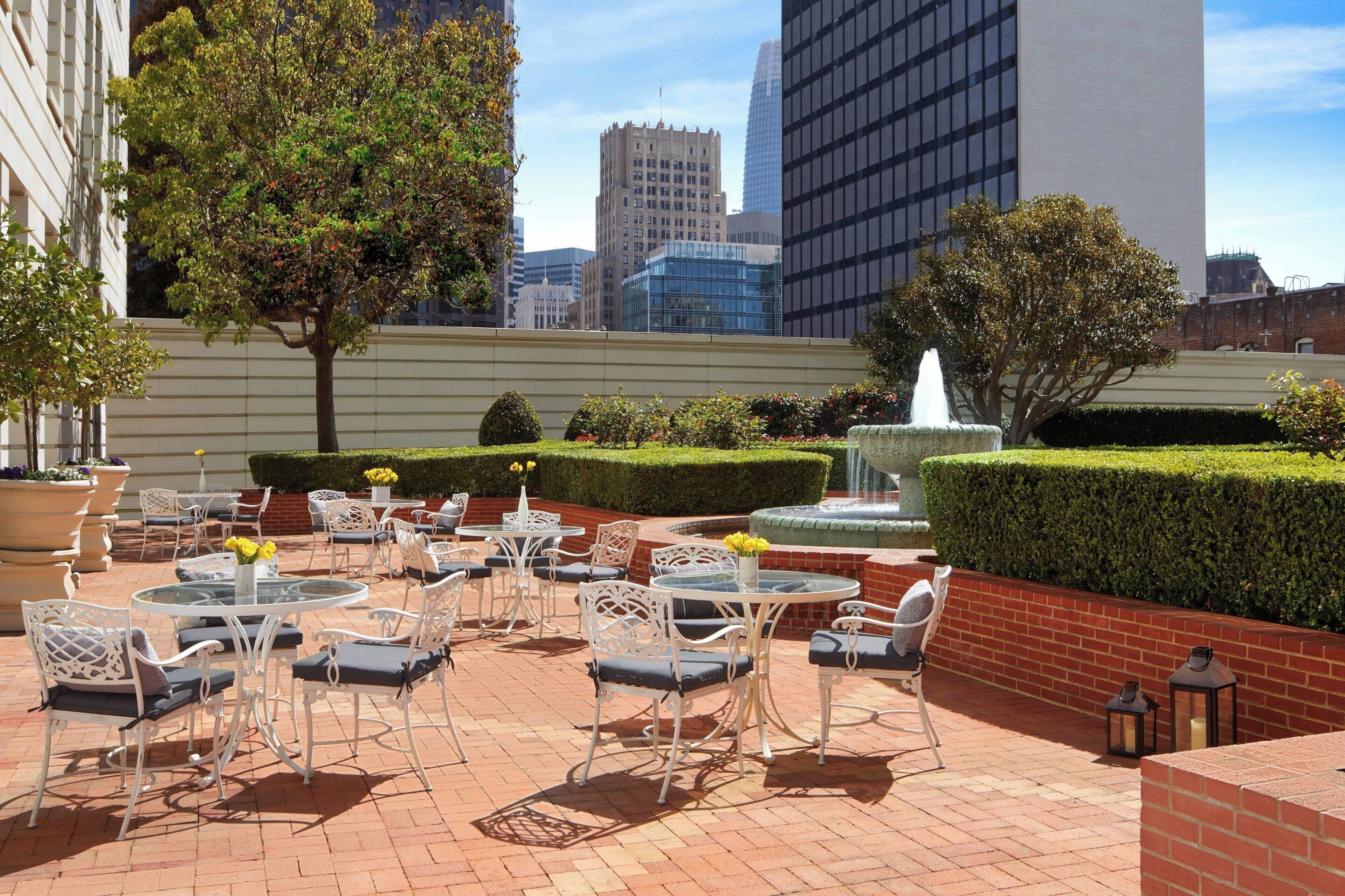 An outdoor patio with round tables and chairs, a fountain, and a view of city buildings.