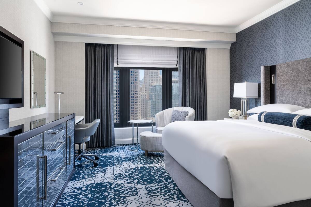 A modern hotel room with a large bed, a desk with a chair, a small table, and a view of the city through the window.