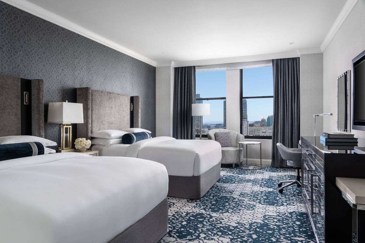 A hotel room with two beds, a desk, a chair, and a large window with a city view.