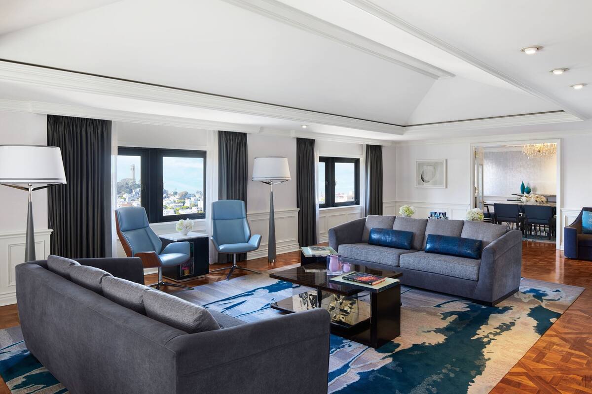 A spacious living room with a grey sofa, blue armchairs, and a coffee table. There are large windows with curtains, a chandelier, and a view of a cityscape.