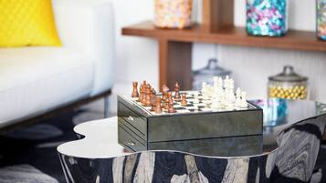 A chess set on a glass table with a modern design, a white sofa with a yellow pillow, and decorative jars on a shelf in the background.