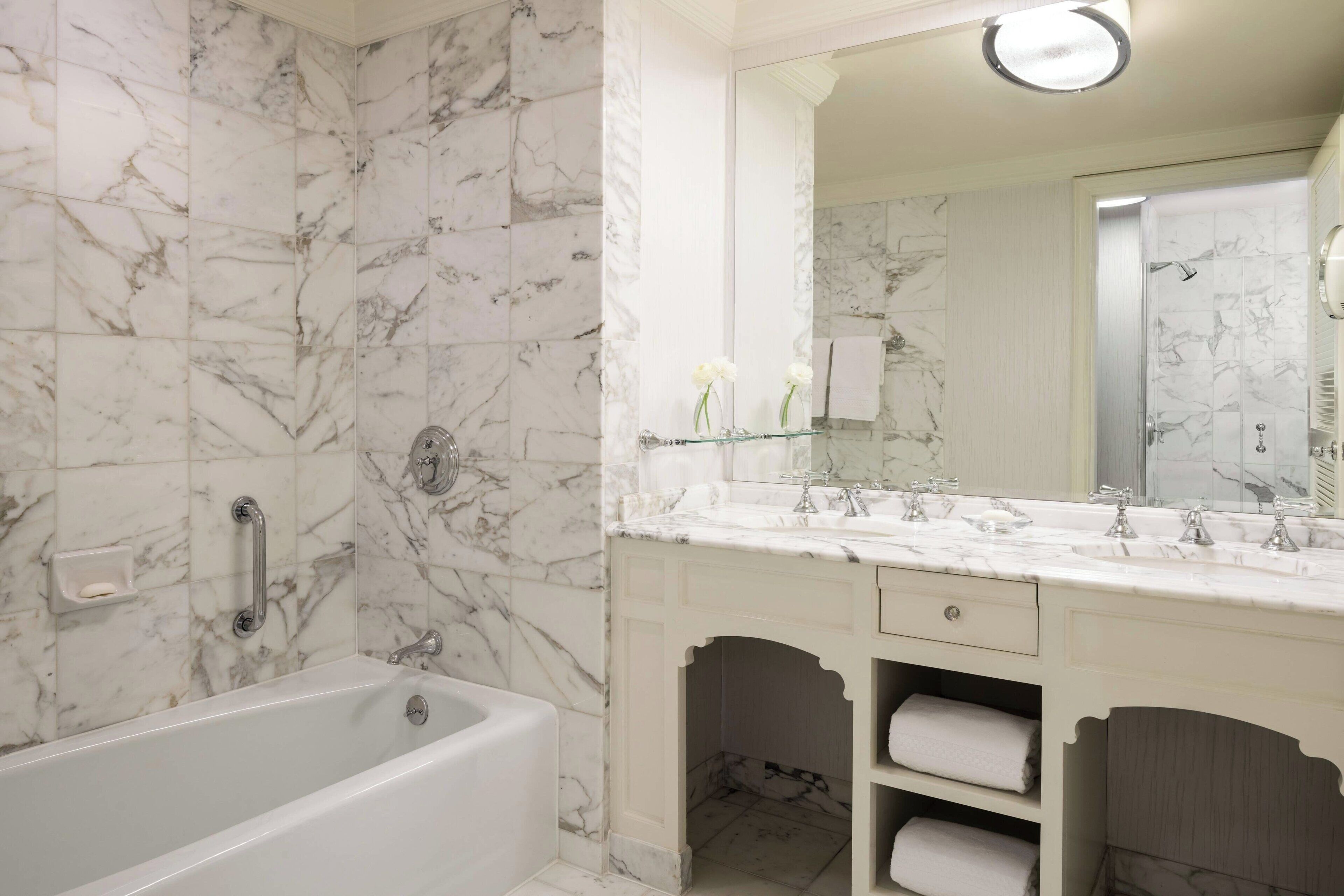 A bathroom with a marble wall, a bathtub, a double sink vanity, and a mirror.