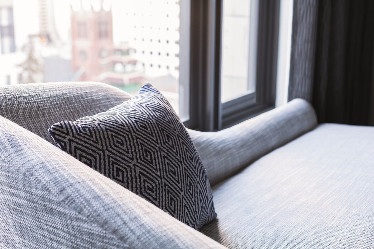 A close-up of a sofa with a patterned pillow and a window with a view of buildings.