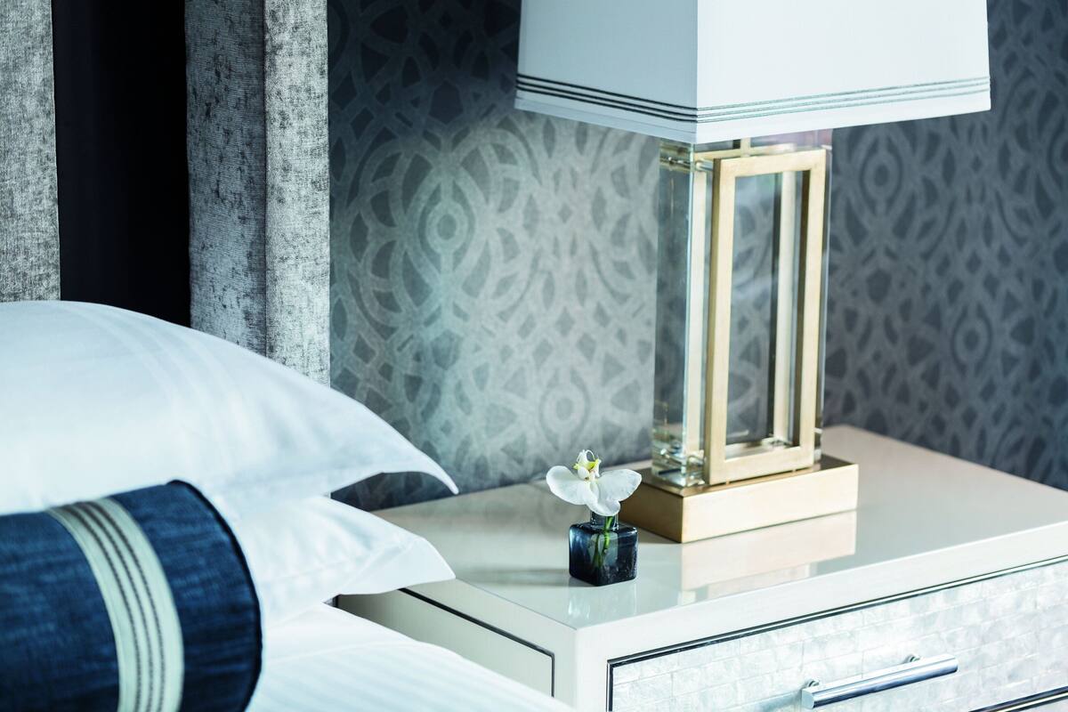A neatly made bed with white linens, a small vase with a single white flower, a bedside table with a lamp, and a patterned wall in the background.