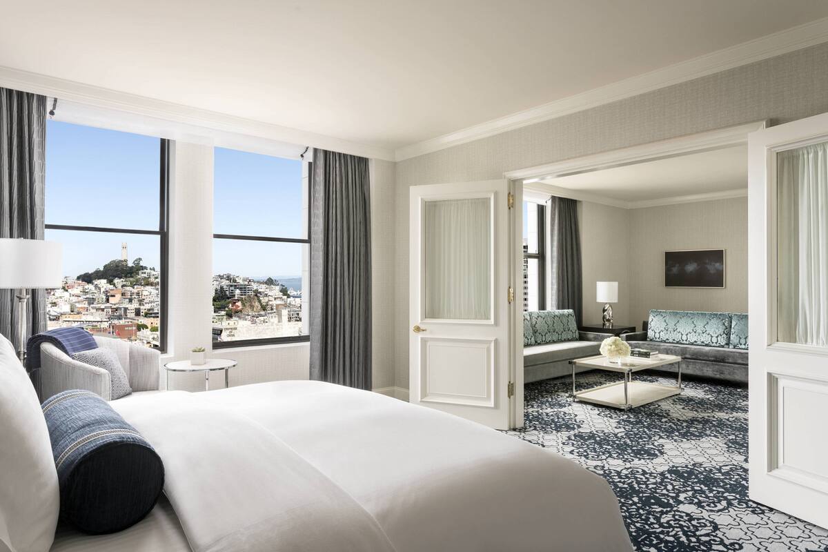 A hotel room with a large bed, a sofa, a coffee table, and a view of a cityscape through large windows.