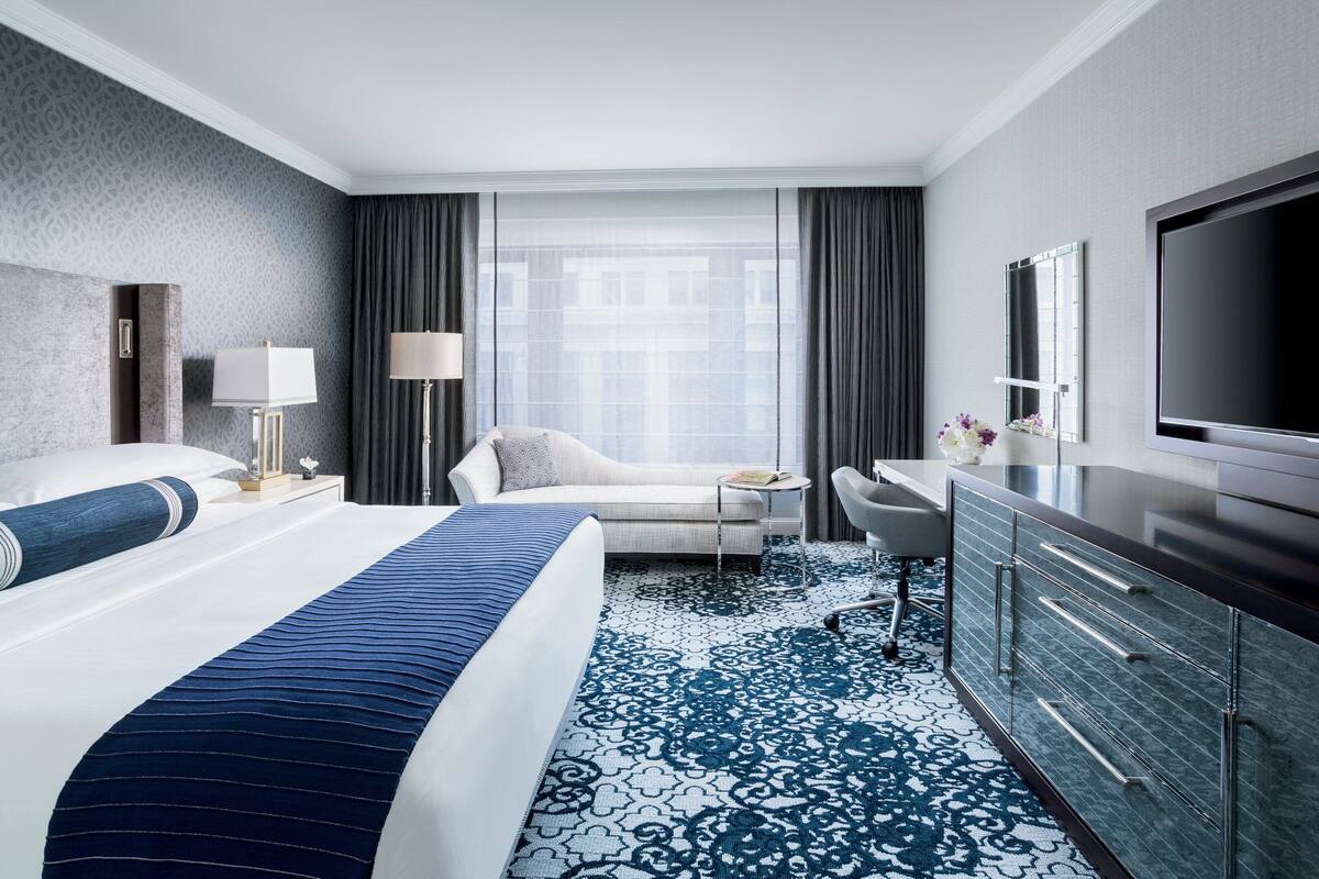 A modern hotel room with a large bed, a desk, a flat-screen TV, and a large window with curtains.